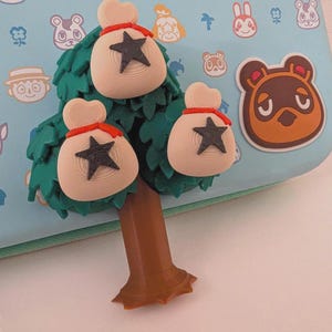 May include: A light blue case featuring various cartoon character designs. A green tree with three beige money bags with black star accents is attached to the case. A brown tree trunk is at the base of the tree.