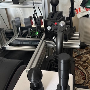 Puede incluir: Un cambio de marchas de simulador de carreras negro con dos botones negros y una palanca negra. El cambio de marchas está montado en una superficie blanca.