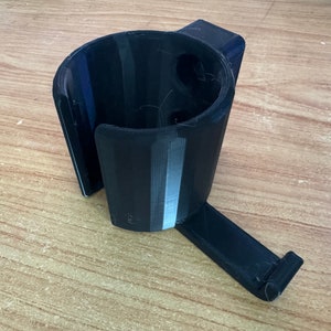 Puede incluir: Un portavasos negro impreso en 3D con un brazo lateral para montaje. El portavasos tiene una superficie texturizada y una abertura circular para un vaso.