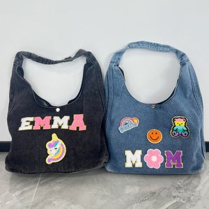 以下が含まれることがあります： カラフルなパッチと刺繍されたテキストが付いたデニムのトートバッグ2つ。1つのバッグには「EMMA」とユニコーンのパッチ、もう1つのバッグには「MOM」と花、スマイリー、クマのパッチが書かれています。