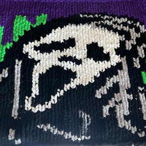 Può includere: Un design a teschio fatto a mano, all'uncinetto in nero, bianco, grigio, verde e viola. Il teschio è il punto focale, con un aspetto dettagliato e strutturato. Lo sfondo è di colore viola.