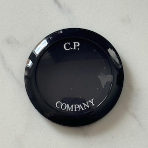 Può includere: Un oggetto nero e circolare con la scritta "C.P." in alto e "COMPANY" in basso in lettere bianche. L'oggetto ha una finitura lucida e sembra essere un coperchio.