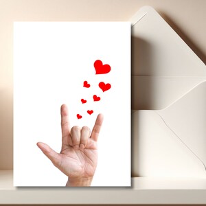 May include: A white card with a red heart and several smaller red hearts falling from the top. The card also features a hand making the American Sign Language sign for "I love you".
