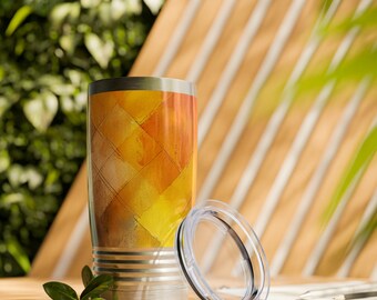 May include: Stainless steel tumbler with a colorful abstract design in shades of yellow, orange, and brown. The tumbler has a silver base and a clear lid. The design features a grid-like pattern. A small green plant is in the foreground.