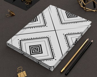 Puede incluir: Un cuaderno blanco con un diseño geométrico negro con cuadrados y líneas punteadas. El cuaderno está sobre una superficie gris oscuro, con clips dorados, lápices y un objeto decorativo dorado cerca.