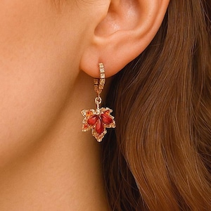 Puede incluir: Pendientes de aro dorados con un diseño colgante de hoja de arce. La hoja presenta piedras preciosas rojas y naranjas, creando una estética otoñal. Los pendientes se muestran sobre un fondo neutro, resaltando los detalles.
