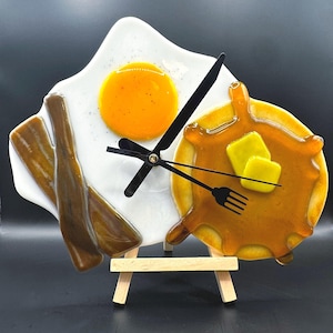 May include: A whimsical breakfast-themed clock featuring a fried egg, bacon, and pancakes with butter and syrup. The clock hands are black, and the clock is mounted on a small wooden easel. The egg yolk is bright yellow, and the bacon is brown.