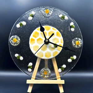 May include: A clear glass clock with a honeycomb design in the center and black clock hands. The clock face features small bee embellishments and is displayed on a wooden easel. The clock is approximately 8 inches in diameter.