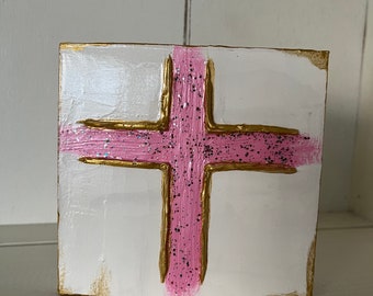 May include: A white canvas with a pink and gold cross painted on it. The cross is outlined in gold and has a pink center with glitter.