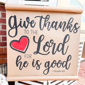 May include: A scroll-style sign with the message "Give Thanks to the Lord for he is good" in black script. A red heart with a black outline is centered on the sign. The sign is made of brown paper and has a wooden dowel at the top and bottom.