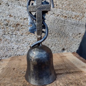 Può includere: Una campana di metallo nero con un design a croce e foglie sulla parte superiore. La campana è appoggiata su una superficie di legno.
