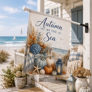 Puede incluir: Una bandera de jardín rectangular con el texto Autumn BY THE Sea. El diseño muestra una escena costera con calabazas azules y naranjas, una hortensia azul, una estrella de mar, una concha y un farol. Una manta a cuadros sobre una silla.