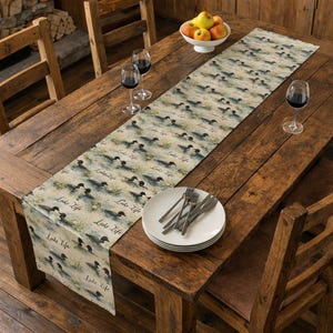 May include: A rustic wooden dining table set with a printed table runner featuring a lake scene with loons and the text "Lake Life." The table is set with plates, silverware, wine glasses, and a bowl of apples.