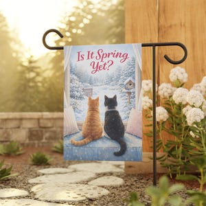 May include: A garden flag featuring a winter scene with two cats looking out a window at a snowy landscape. The flag reads "Is It Spring Yet?" in pink script. The cats are orange and black, and the flag is displayed on a black metal stand.