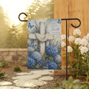 Puede incluir: Una bandera de jardín con una cruz blanca cubierta con una tela blanca, rodeada de hortensias azules y mariposas. La bandera tiene el texto "I WILL WALK BY Faith EVEN WHEN I CANNOT SEE."