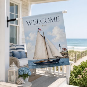 May include: A rectangular garden flag featuring the word WELCOME in dark blue text at the top. The image depicts a dark blue sailboat with white sails on the water, a small American flag on the mast, and a lighthouse on the rocky shore.
