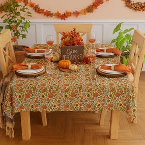 May include: A Thanksgiving table setting with a floral tablecloth featuring pumpkins and sunflowers. The table is set with plates, napkins, and wine glasses. A wooden sign reads "Give Thanks". Decorative gourds and fall foliage complete the festive scene.