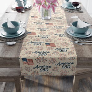 May include: A beige table runner with a repeating pattern of the American flag, fireworks, and the text "America 250" in navy blue and red. The runner is set on a wooden table with place settings.