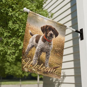 Op de afbeelding: Een decoratieve buiten vlag met een bruine en witte puppy met een rode halsband en een kleine bel. De vlag heeft het woord "Welcome" in een cursief lettertype. De achtergrond toont een veld met hoog gras.
