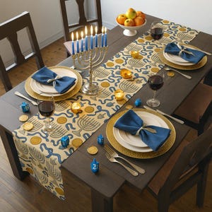 May include: A festive table setting for Hanukkah. The dark wood table is set with gold-rimmed plates, blue napkins, and silverware. A menorah with lit candles sits in the center, along with a patterned table runner, dreidels, and wine glasses. A bowl of fruit is in the background.