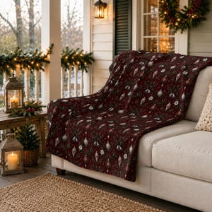 May include: A dark red throw blanket featuring a repeating pattern of silver Christmas ornaments, bells, and white flowers. The fabric has a soft texture and is draped over a light-coloured sofa, showcasing a festive holiday-themed design.