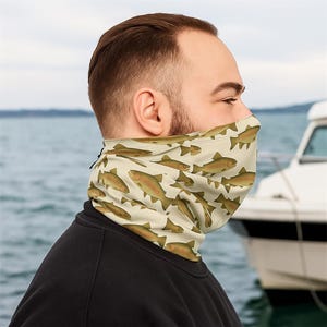 Könnte beinhalten: Ein beigefarbener Halstuch mit einem sich wiederholenden Muster aus braunen Fischen. Das Halstuch wird um den Hals getragen und bedeckt das untere Gesicht. Im Hintergrund ist ein Boot auf dem Wasser zu sehen.