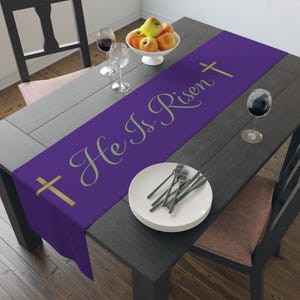 May include: A purple table runner with the text "He Is Risen" in a script font, along with gold crosses. The runner is placed on a dark wooden table, set with plates, silverware, and wine glasses, and a bowl of apples.