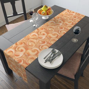 May include: A dining table set with a decorative orange and cream swirl patterned table runner. The table is set with white plates, silverware, wine glasses, and a bowl of apples. The table and chairs are dark gray.