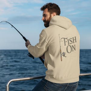May include: A beige hoodie with the text "Fish On" and a fishing rod graphic on the back. The person in the image is wearing blue jeans and holding a fishing rod, with the ocean and sky in the background.