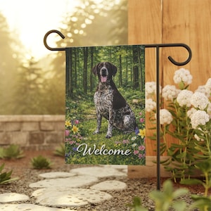 May include: A garden flag featuring a German Shorthaired Pointer dog sitting in a forest setting. The flag has the word "Welcome" in script. The dog is brown and white, and the background includes trees and flowers.