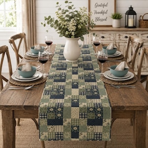 May include: A rectangular table runner featuring a patchwork design in shades of navy blue, sage green, and cream. The pattern consists of alternating squares and rectangles with floral, plaid, and solid prints, laid flat on a wooden dining table.