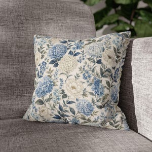 Könnte beinhalten: Ein quadratisches Zierkissen mit einem Blumenmuster aus blauen Hortensienclustern, weißen pfingstrosenartigen Blüten und grünem Blattwerk auf cremefarbenem Grund. Der Stoff weist eine deutlich sichtbare Webstruktur auf der gesamten Vorderseite auf.