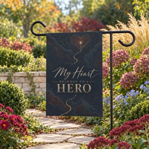 May include: A rectangular garden flag hanging on a black metal stand. The flag features a dark navy background with topographic lines, a gold star, and a heart shape. It displays the text "My Heart BELONGS TO A HERO" in white and gold.