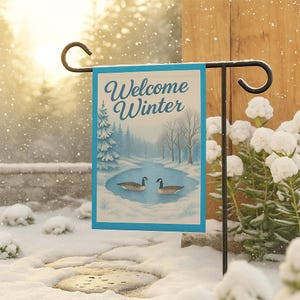 May include: A decorative garden flag with a winter scene. The flag features a blue border and the words "Welcome Winter" in script. The image depicts a snowy landscape with a pond and two geese. The flag is hanging on a black metal stand.
