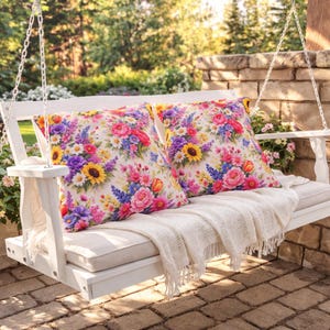 May include: A white wooden garden swing with two cushions adorned with a colourful floral pattern. The cushions display sunflowers, roses, and other flowers in pink, yellow, purple, and blue. A cream-coloured throw blanket and white chains complete the scene.
