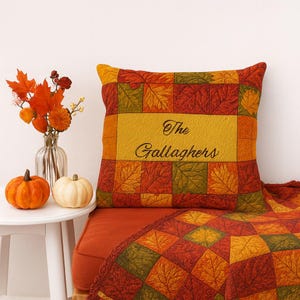 Op de afbeelding: Een decoratief vierkant kussen met een herfstbladerenpatroon in tinten rood, oranje en groen. Het kussen heeft een geel middenpaneel met de tekst "The Gallaghers" in zwart script. Een bijpassende deken is over een stoel gedrapeerd.