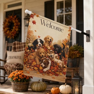 May include: A rectangular garden flag featuring the text Welcome at the top and an illustration of six puppies of various breeds playing in a pile of orange and red autumn leaves. The flag is mounted on a white pole attached to a black wall bracket.