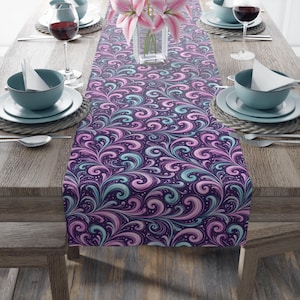 May include: A decorative table runner with a swirling, floral pattern in shades of purple, pink, and teal. The runner is placed on a wooden table set for a meal, with plates, bowls, and wine glasses.