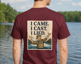 May include: Maroon t-shirt with a humorous fishing-themed graphic on the back. The graphic features a cartoon of a fisherman in water with the text "I CAME. I CAST. I LIED." The shirt is worn by a person standing in front of a lake.