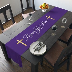 May include: A purple table runner with the text "Prepare Your Heart" and gold crosses. The runner is placed on a dark wooden table set for a meal, with plates, silverware, and wine glasses. A bowl of fruit sits nearby.