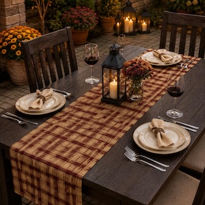 May include: A rectangular table runner featuring a brown and tan plaid pattern, displayed on a dark wooden dining table set with cream-colored plates, beige napkins with dark rings, silver forks, and wine glasses, centered by a black lantern.