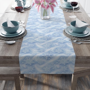 May include: A light blue table runner with a white snowflake pattern. The runner is placed on a wooden table set for a meal, with plates, bowls, silverware, and wine glasses. A vase of pink flowers is in the center.