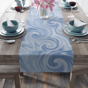 May include: A blue and white swirl patterned table runner. The runner is placed on a wooden table set for a meal, with plates, bowls, silverware, and wine glasses. A vase of pink flowers sits in the center.