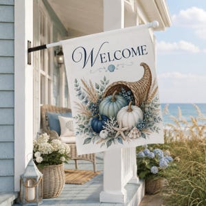 Puede incluir: Una bandera de jardín rectangular con la palabra Welcome en letra cursiva azul oscuro en la parte superior. El diseño muestra un cuerno de la abundancia tejido lleno de calabazas azul claro, azul oscuro y blancas, trigo seco, hojas de eucalipto, una estrella de mar y conchas.