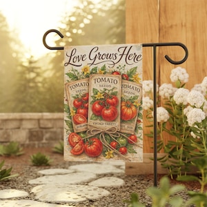 May include: A garden flag with the text "Love Grows Here" and illustrations of tomato seed packets and ripe tomatoes. The flag is displayed on a black metal stand in a garden setting.
