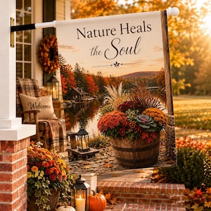 May include: A rectangular garden flag featuring the text Nature Heals the Soul above a scenic autumn landscape. The image displays a wooden barrel filled with colourful orange and red chrysanthemums, a lake, trees, and a small cabin at sunset.