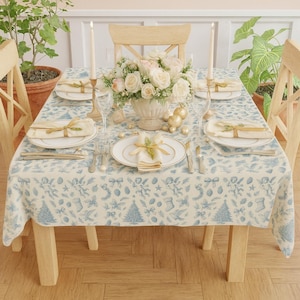 以下が含まれることがあります： 青いホリデー柄のクリーム色のテーブルクロスが敷かれたお祝いのテーブルセッティング。白いお皿、金縁のカトラリー、リネンのナプキンがセットされています。フラワーアレンジメントとキャンドルが、エレガントなホリデーデコレーションを完成させています。