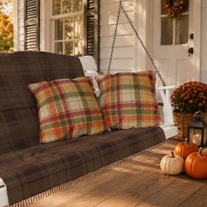 May include: A white wooden porch swing featuring a dark brown plaid throw blanket and two square pillows with a multicolored plaid pattern in shades of orange, yellow, green, and red. The swing is suspended by metal chains on a wooden porch.