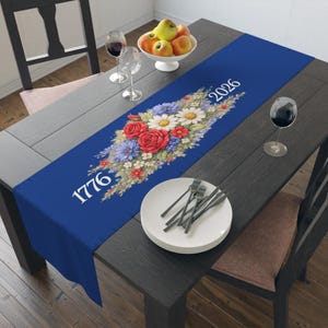 Puede incluir: Camino de mesa azul con un diseño floral con rosas rojas, margaritas blancas y flores azules. Los números "1776" y "2026" están impresos en blanco. Colocado sobre una mesa de madera oscura.