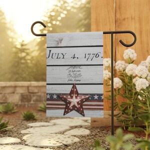 May include: A garden flag with a patriotic theme. The flag displays the text "July 4, 1776" and historical figures' names. A red star with white stars is centered on a ribbon with red and blue stripes and white stars.